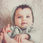 A cute baby in a knitted outfit lying on a soft carpet, smiling warmly.